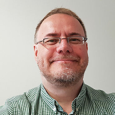 Selbstporträt von Matthias Rillig. Er trägt kurze, Blonde Haare, einen kurzen Bart, eine eckige randlose Brille und ein kariertes Hemd.
