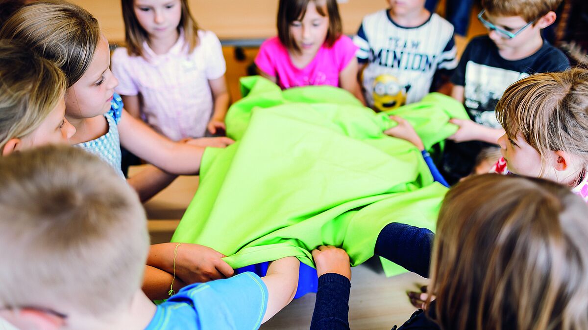 Auf dem Bild sieht man viele Kinder, die gemeinsam ihre Hände unter einem grünen Tuch halten