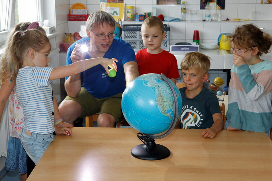 Ein Mann und fünf Kinder sitzen um einen Tisch auf dem ein Globus steht. Der Mann leuchtet mit einer Taschenlampe. Ein Kind hält eine Kugel.