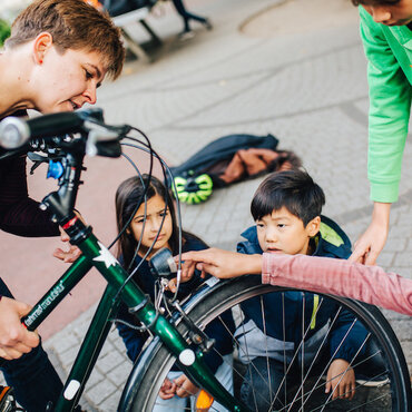 Eine Lernbegleitung erforscht auf dem Schulhof mit vier Kindern im Grundschulalter ein Fahrrad. Gemeinsam betrachten sie die Frontleuchte des Rads.