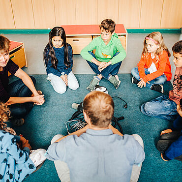Eine Pädagogin und ein Pädagoge sitzen mit Kindern im Kreis auf dem Boden und sind im Gespräch.