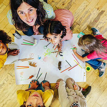 Eine Erzieherin sitzt mit Kindern auf dem Boden und malt mit Buntsfiften auf einem Blatt Papier
