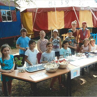 Eine Gruppe von Kindern und zwei Erwachsenen stehen und sitzen hinter einem langen Tisch und präsentieren selbstgemachte Brötchen und Marmelade.